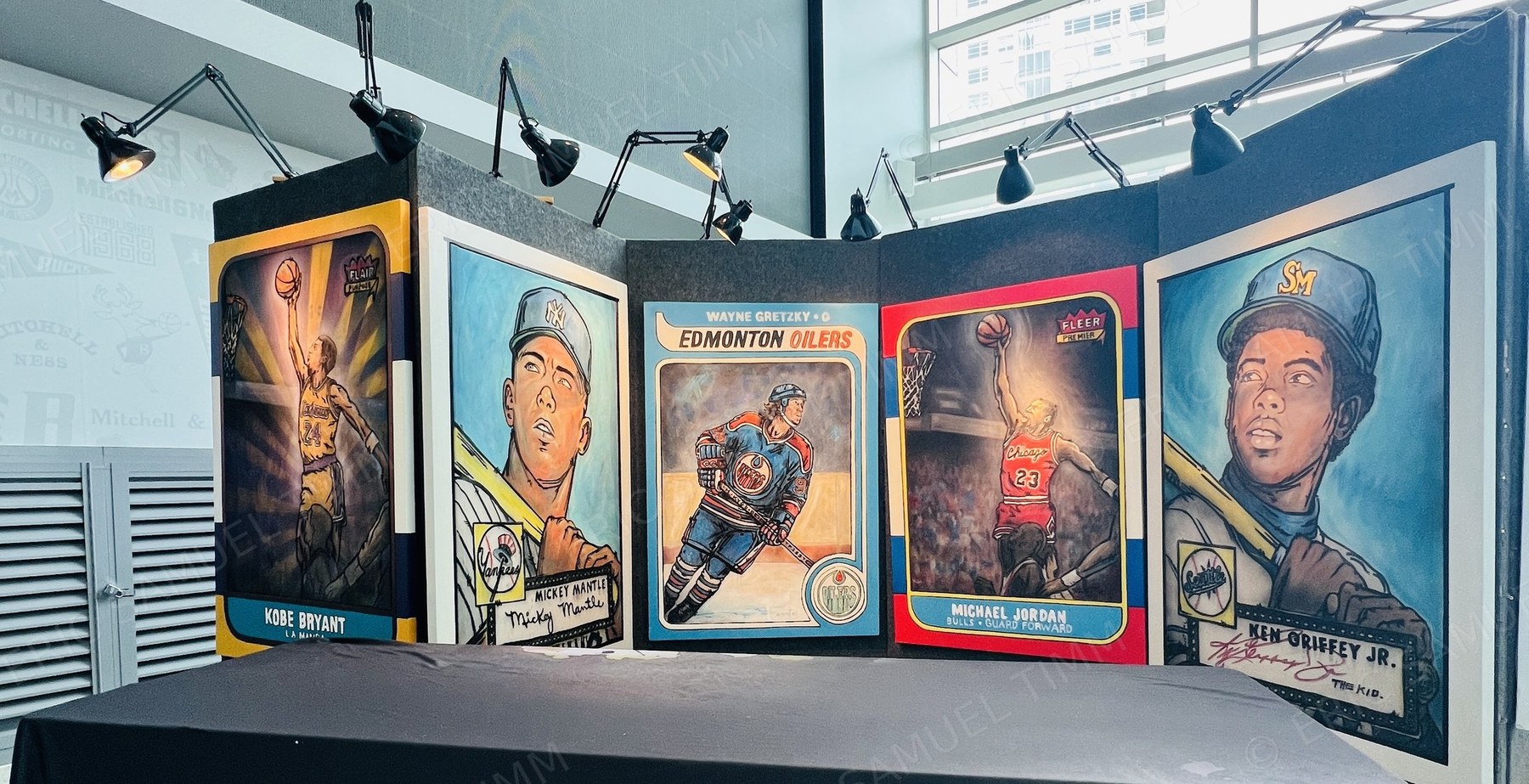 Five original paintings — Kobe Bryant, Mickey Mantle, Wayne Gretzky, Michael Jordan and Ken Griffey Jr. — standing at full height on a lit display wall behind a table